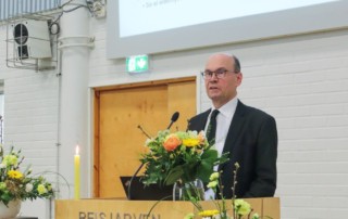 Ari-Pekka Palola esitelmöimässä Reisjärven opistolla. Palola seisoo puisen puhujankorokkeen takana. Taustalla on diaesitys aiheesta.